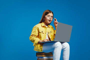 Astonished girl with glasses and a yellow jacket engrossed in using a laptop, seated on a stack of books, capturing a sense of curiosity and learning. Ideal for educational content