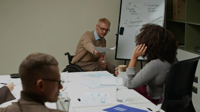 Male office manager using wheelchair looking through document then passing printout to colleague rejecting idea and continuing presentation on workflow strategy during meeting in office
