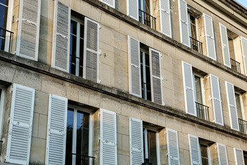 Obraz premium A detailed view of the upper floors of a traditional Parisian stone building, emphasizing the repeating pattern of windows with classic white wooden shutters. 