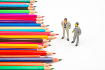 Colorful pencils spread across a white surface, with miniature businessmen standing, merging...