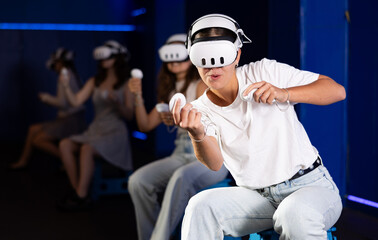 Cheerful emotional young female in VR goggles sitting in virtual reality room with group of friends, manipulating objects or shooting at targets with gaming controllers in hands.