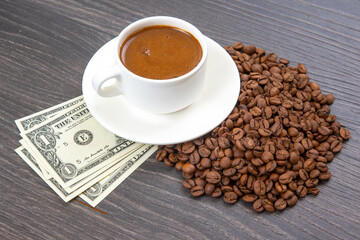 White cup of freshly brewed coffee on a saucer, surrounded by roasted coffee beans and dollar bills, placed on a dark wooden surface.