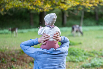 Rear view of a father lovingly holding his baby on his shoulders and watching deers in the background. Natural green surroundings in an open forest area.