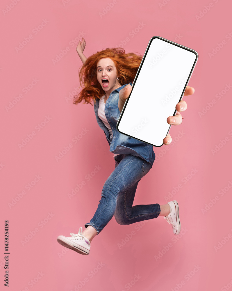 Poster excited redhead woman having fun, jumping,demonstrating mobile phone with empty white screen on pink