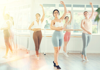 Naklejka premium Expressive young dark-haired woman in twenties, wearing workout clothes, mastering ballet moves at group class in sunlit dance studio
