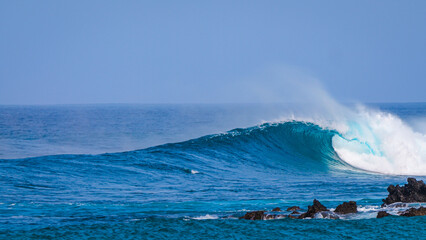 Perfect glassy wave curls and breaks gracefully near a jagged volcanic shoreline. Turquoise ocean glistens and creates a surfable wave framed by rocky outcrops of the coastline at Canary Islands.