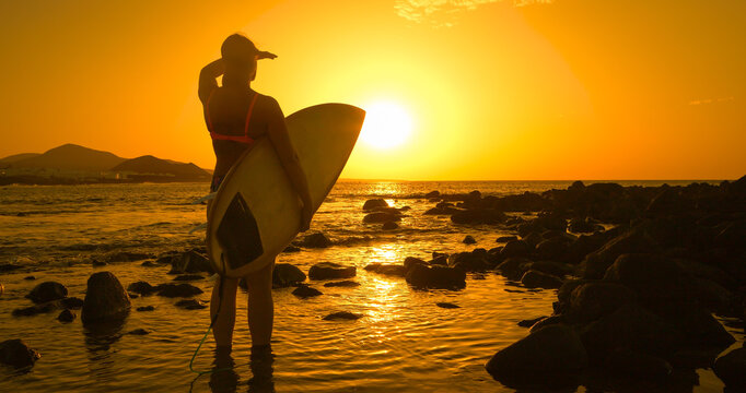 SILHOUETTE, LENS FLARE: Female surfer stands in shallow water, shielding her eyes as she looks out to sea at golden sunset. She is checking out the surf spot before heading for an evening session.
