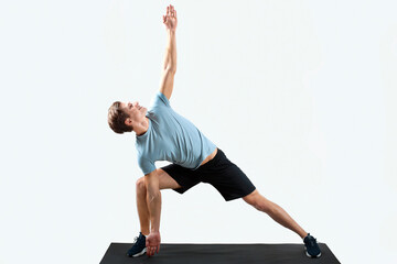 Strong young man standing in yoga pose on sports mat over light studio background, full length...