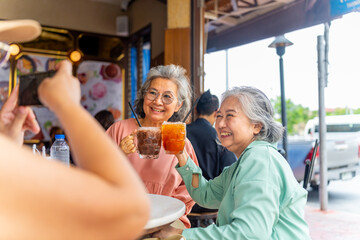 Happy Asian senior woman enjoy travel in the city on summer holiday vacation. Elderly women friends using smartphone taking picture during travel street market and eating street food at outdoor cafe.