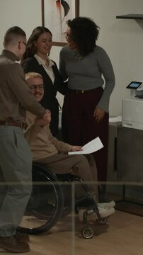 Vertical full shot of cheerful corporate workers meeting at photocopier in office hallway greeting each other and chatting amicably at workplace