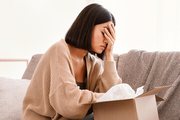 Wrong Item Concept. Sad unpset asian lady sitting on couch and unpacking cardboard box, received...
