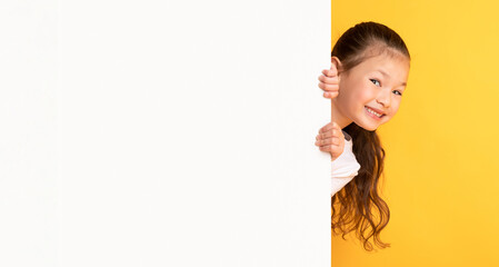 Advertisement And Sale Concept. Portrait of playful asian child peeking out blank white poster with mock up template and free space for logo, design or message. Cute girl hiding behind placard