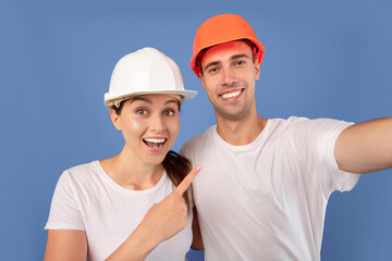 Repair Home Concept. Close up of young couple in protective helmets taking selfie together during renovation, cheerful man and woman in hard hats smiling at camera over blue background, free space