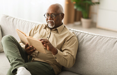 Mature Black Man Reading A Paper Book Relaxing Sitting On Sofa At Home, Wearing Glasses. Senior...