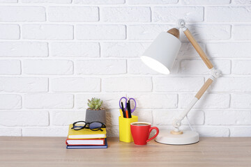 Different stationery, coffee, lamp, glasses and floral decor on wooden office table near white brick wall