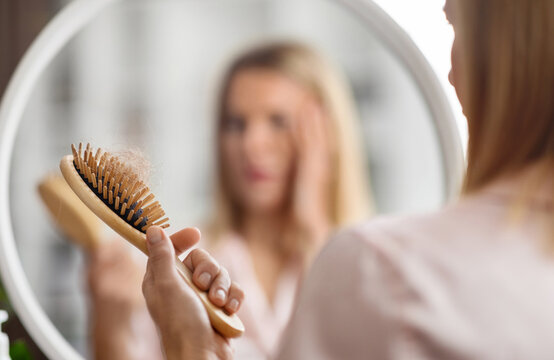 Hairloss Concept. Shocked Blonde Woman Holding Comb Full Of Fallen Hair After Brushing, Closeup Shot Of Middle Aged Lady With Brush In Hand Standing Near Mirror, Suffering Alopecia Or Health Problems