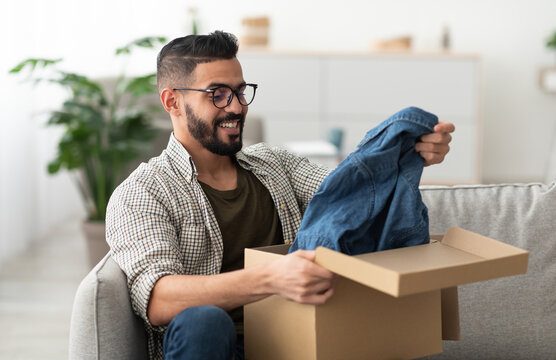 Satisfied Arab male client taking out new clothes from cardboard box, happy with delivery service at home. Millennial Eastern guy unpacking cardboard parcel, receiving online store order, indoors