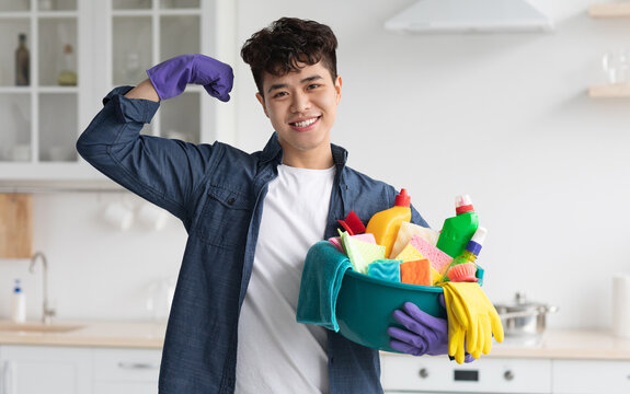 Positive asian guy with cleaning supplies showing his muscles and smiling at camera. Enthusiastic chinese man wearing gloves, holding busket with sprays and dust clothes, cleaning kitchen, free space