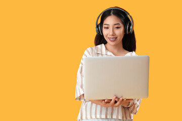 Happy young female Asian programmer in headphones with laptop on yellow background