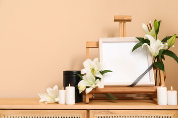 Blank funeral frame with lily flowers, mortuary urn and burning candles on wooden cabinet against...