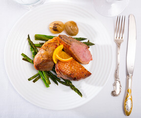 Top view of juicy roasted duck breast served on white plate with baked onions, pickled asparagus and slice of orange