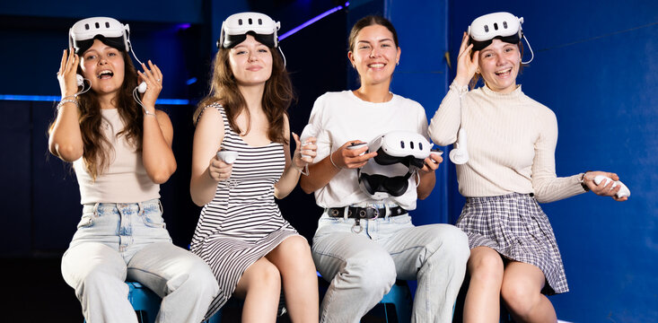 Group of young girls with VR headsets and controllers, eagerly anticipating virtual reality experience during fun-filled get-together, sitting on stools in modern gaming lounge