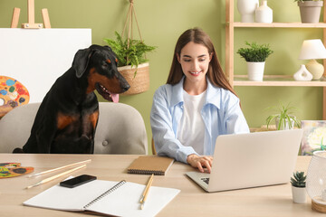 Cute Doberman dog near paints palette and beautiful young happy woman working with laptop at home