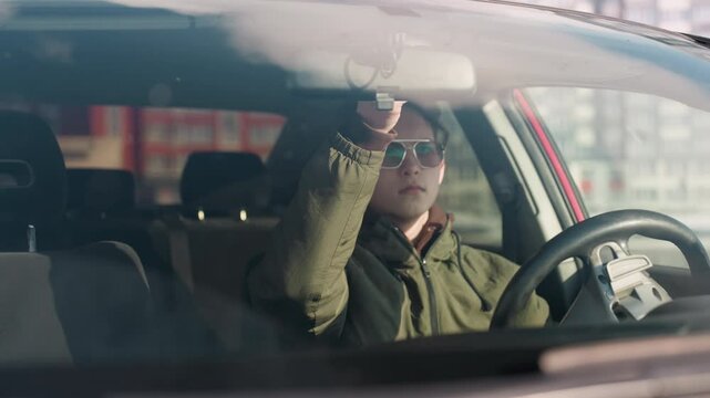 young man wearing winter jacket in parked car lifts sun visor upward while seated in driver seat on bright day with reflections on windshield and urban buildings visible in background