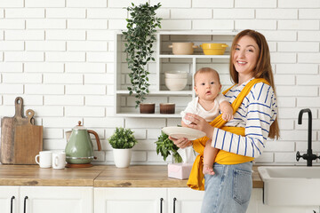 Young mother with little baby in sling wiping plate at home