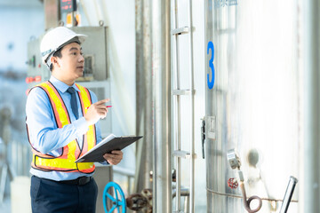 Fototapeta premium Engineer or inspector wearing a safety helmet and reflective vest, checking equipment and taking notes at an industrial plant. Safety and quality control concept.