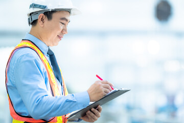 Smiling engineer in a safety helmet and reflective vest checking documents at an industrial plant....