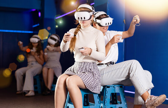 Cheerful young female friends wearing VR headsets and holding controllers having fun in virtual reality studio, competing in two-on-two shooter game