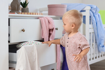 Naughty baby boy opening drawer with clothes in messy bedroom