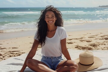 Smiling woman sitting cross-legged on the beach. She is wearing a white t-shirt and jean shorts. Beach hat on the ground next to her.