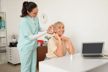 Doctor putting hearing aid on mature woman's ear in clinic