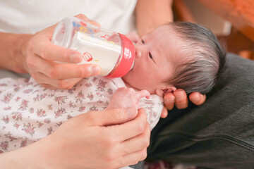 A 7-day-old baby drinks milk from a bottle while lying on the lap of a Japanese dad in his 20s. This tender moment shows quiet love and the start of fatherhood. Great for parenting or family content.