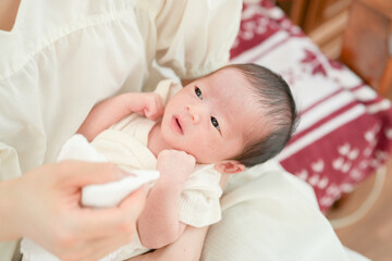A Japanese mother in her twenties holds her 7-day-old newborn baby and wipes the baby's mouth while sitting on a sofa. This image shows the love and care shared in the first days of motherhood.