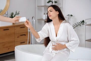 Young woman with menstrual cramps taking estrogen pills from person in bathroom