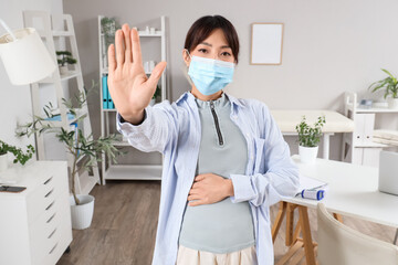 Pregnant Asian woman in medical mask showing STOP gesture at hospital