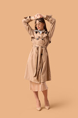 Pretty young woman in stylish trench coat and sunglasses on beige background