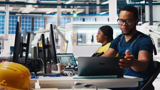 Black engineer inputs solar system parameters at industrial workstation, using PC in a renewable energy facility. Technician supports sustainable production through software management. Camera B.