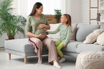 Obraz premium Beautiful young happy pregnant woman with her cute little daughter sitting on sofa at home