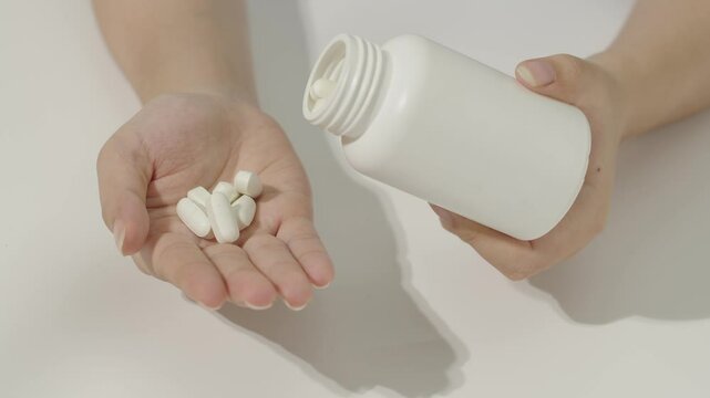 Close-up of hand holding pill bottle opening and pouring white pills into hand below. The compressed pills have a border in the middle to make it easy to break in half. Presented on white background.