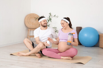Sporty pregnant couple with pill bottle and water bottle sitting on mat at gym