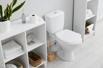 Ceramic toilet bowl, brush and shelving units with bath supplies near light wall
