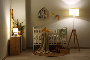 Interior of children's bedroom with baby crib, dresser and glowing lamps near white wall at night