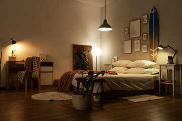Interior of dark bedroom with surfboard, tables and glowing lamps in evening