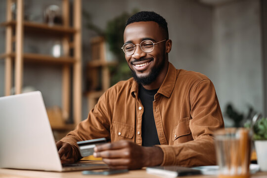 Happy african american businessman is making online payment holding credit card, using laptop, shopping online at workplace