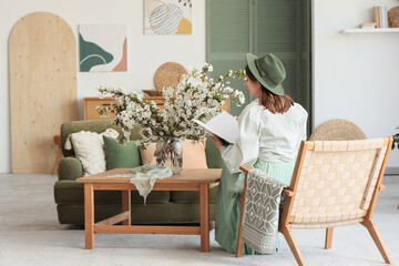 Young woman on armchair and vase with flowering branches in living room, back view