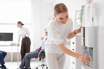 Fototapeta premium Young businesswoman pouring water from cooler in office